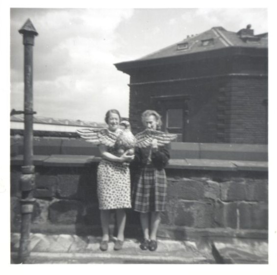 A black and white photograph of two women in patterned dresses standing on a roof. In their arms, they are holding a large carved and gilded wooden eagle. Behind them is a brick building and part of a train station.