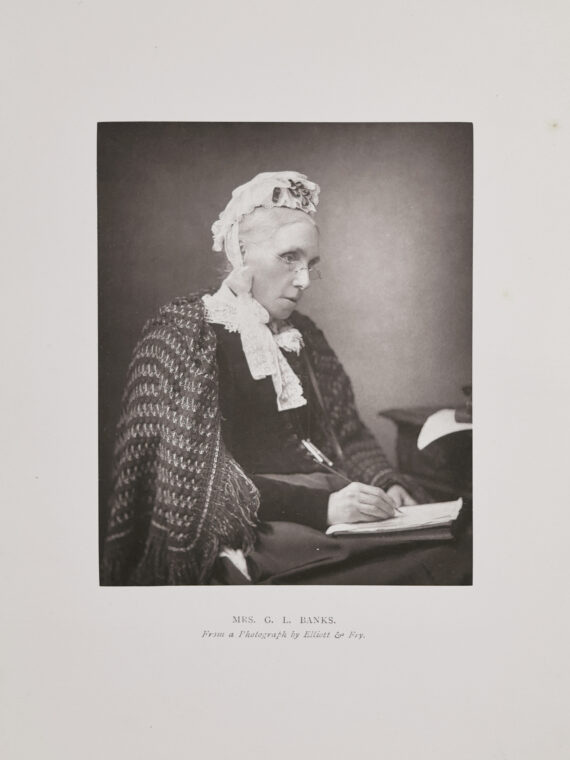 A photograph of an older Isabella Banks. She is wearing glasses, a cap and a shawl, and sits with a pen and paper in her hands.