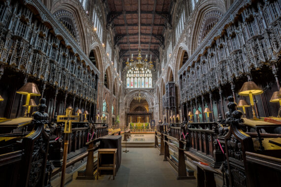 The quire of Manchester Cathedral, looking upwards. On either side of the centre of the quire there are intricately-carved wooden quire stalls, and the ceiling is comprised of carved wooden panels. In the distance is the altar. 