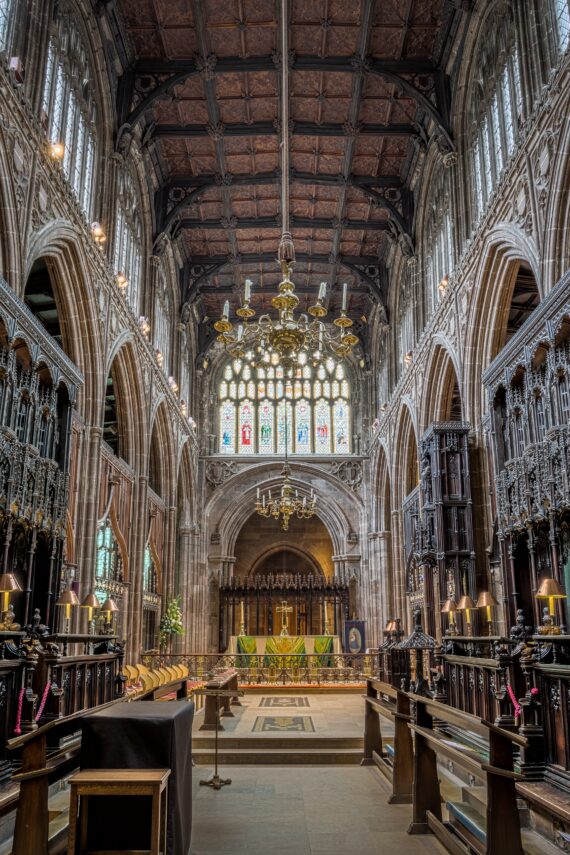 The quire of Manchester Cathedral, looking upwards. On either side of the centre of the quire there are intricately-carved wooden quire stalls, and the ceiling is comprised of carved wooden panels. In the distance is the altar. 