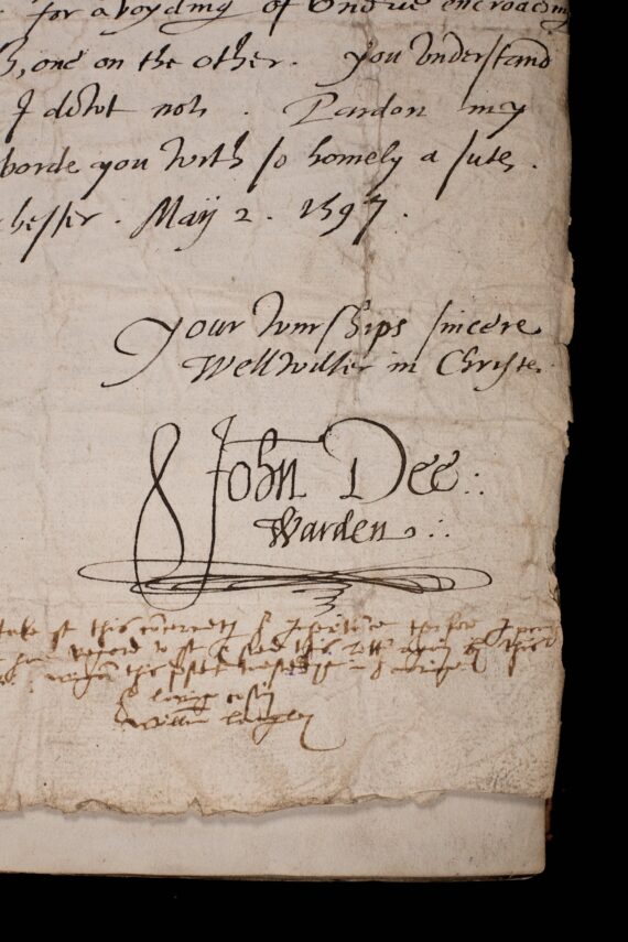 The corner of a sheet of sheet of paper containing a letter. A cursive hand has written ‘Your wurships sincere wellwisher in Christe’, and beneath it written ‘John Dee: Warden’.