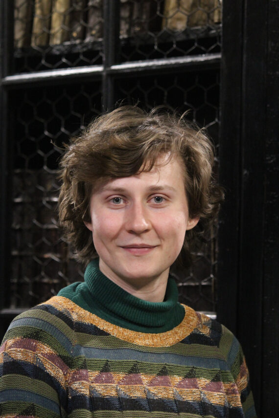 A clean-shaven man with scruffy brown hair and no glasses, wearing a green turtleneck sweater and a green patterned knitted jumper over it.