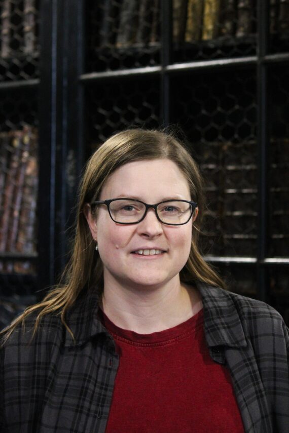 A woman with straight brown hair and black-rimmed glasses, wearing a red t-shirt and a chequered grey shirt over it.