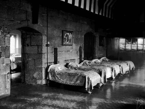 A row of single beds covered with blankets stand with their heads against a medieval stone wall. Through a ‘Tudor’ arch, another room containing more beds and a medieval arched window is visible. Above, a medieval timbered roof is visible.