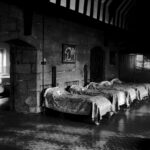 A row of single beds covered with blankets stand with their heads against a medieval stone wall. Through a ‘Tudor’ arch, another room containing more beds and a medieval arched window is visible. Above, a medieval timbered roof is visible.