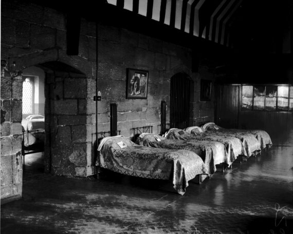 A row of single beds covered with blankets stand with their heads against a medieval stone wall. Through a ‘Tudor’ arch, another room containing more beds and a medieval arched window is visible. Above, a medieval timbered roof is visible.