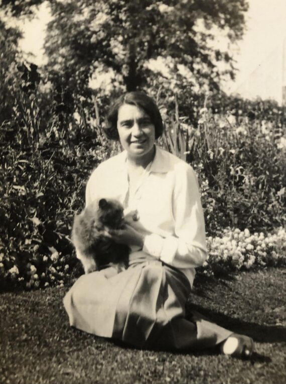 A black and white photograph of a woman wearing a white shirt and a skirt and sitting on the grass in front of a flower bed. She holds a cat in her arms, and is looking at the camera.