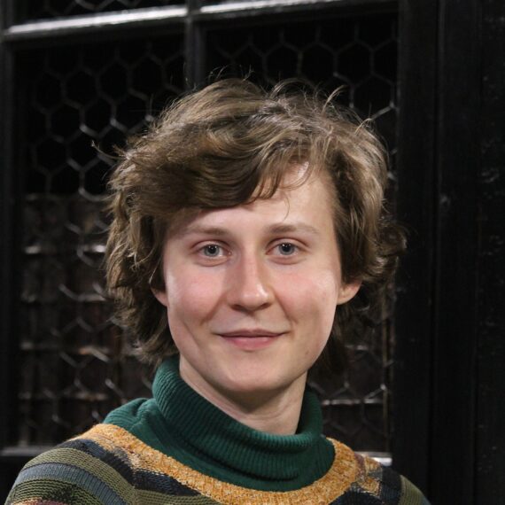 A clean-shaven man with scruffy brown hair and no glasses, wearing a green turtleneck sweater and a green patterned knitted jumper over it.