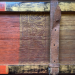 A partial image of the painted foredge of a book, the pages of which are made from palm leaves. To the left (the centre of the foredge) the palm leaves are painted read, while to the right, they are gold. A floral design in red against a gold background separates the two sections. The wooden covers are similarly coloured, and are held together by a leather strap with holes and a buckle.