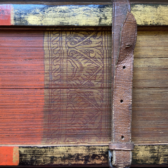 A partial image of the painted foredge of a book, the pages of which are made from palm leaves. To the left (the centre of the foredge) the palm leaves are painted read, while to the right, they are gold. A floral design in red against a gold background separates the two sections. The wooden covers are similarly coloured, and are held together by a leather strap with holes and a buckle.