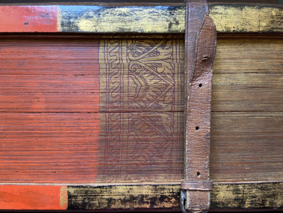 A partial image of the painted foredge of a book, the pages of which are made from palm leaves. To the left (the centre of the foredge) the palm leaves are painted read, while to the right, they are gold. A floral design in red against a gold background separates the two sections. The wooden covers are similarly coloured, and are held together by a leather strap with holes and a buckle.