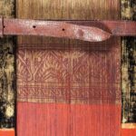 A partial image of the painted foredge of a book, the pages of which are made from palm leaves. At the bottom (the centre of the foredge) the palm leaves are painted read, while at the top, they are gold. A floral design in red against a gold background separates the two sections. The wooden covers are similarly coloured, and are held together by a leather strap with holes and a buckle.
