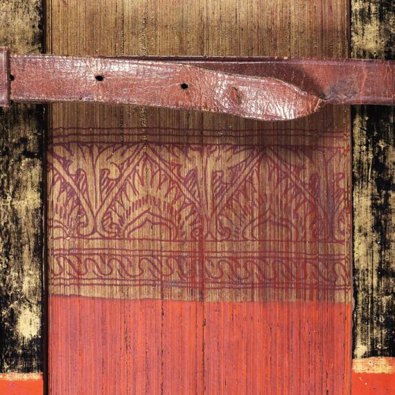 A partial image of the painted foredge of a book, the pages of which are made from palm leaves. At the bottom (the centre of the foredge) the palm leaves are painted read, while at the top, they are gold. A floral design in red against a gold background separates the two sections. The wooden covers are similarly coloured, and are held together by a leather strap with holes and a buckle.