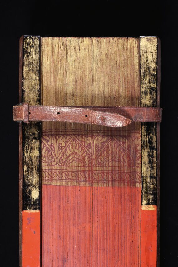 A partial image of the painted foredge of a book, the pages of which are made from palm leaves. At the bottom (the centre of the foredge) the palm leaves are painted read, while at the top, they are gold. A floral design in red against a gold background separates the two sections. The wooden covers are similarly coloured, and are held together by a leather strap with holes and a buckle.