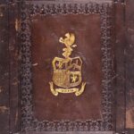A close-up of the cover of a book bound in dark brown leather, now quite scuffed. In the centre of the cover is a gilt-stamped armorial design featuring the arms of Humphrey Chetham, and beneath it, a scroll bearing the motto ‘QUOD TUUM TENE’. Around the edges is a blind-stamped decorative border.