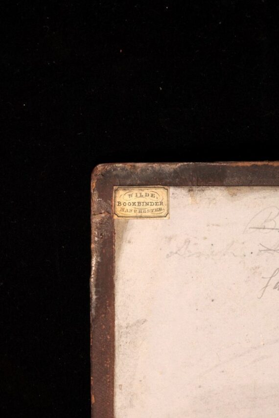 The upper left corner of the inside of a book’s front cover. A blank page has been pasted down to the brown leather turn-ins of the cover, and in the very upper left corner is a small pasted rectangle, on which is printed (on three lines) ‘WILDE. BOOKBINDER. MANCHESTER’. 