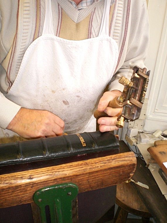 The torso of a man wearing an apron is visible behind a wooden and metal stand holding a book bound in black leather. The words ‘Holy Bible’ have been embossed in gold on the spine of the book by the man, using a tool that he holds in his left hand.