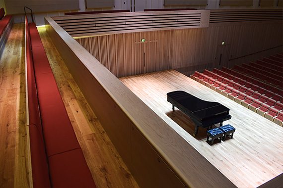 The Stoller Hall's bench gallery seating, which has red fabric cushioning (left) with a view down to the grand piano on the stage (right)