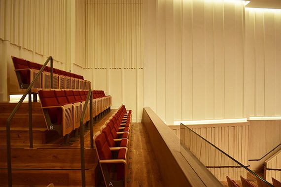 A side-on image of The Stoller Hall's balcony, seating (left). The seats are pale wood, covered in red fabric cushioning.