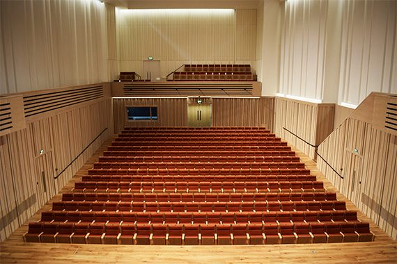 A view of The Stoller Hall's stall and balcony seating, which is straight rows of red fabric cushioned chairs with pale wood armrests.