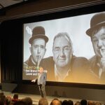 Neil Brand speaks in front of a cinema screen showing Laurel and Hardy