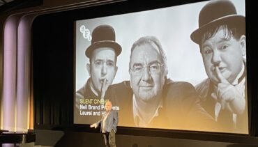 Neil Brand speaks in front of a cinema screen showing Laurel and Hardy