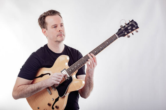 Jim Faulkner with pale wood guitar on pale grey background
