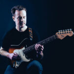 Jim Faulkner with electric guitar, on dark blue background