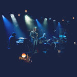 Singer Kurt Elling (holding a tambourine) with keyboard player (left) and guitarist (right) against blue lighting