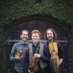 Folk/Classical group Lodestar Trio, holding stringed instruments, against a dark green backdrop