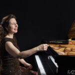 Pianist Angela Hewitt sits at a grand piano, wearing a brown dress