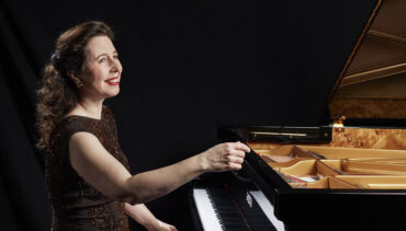 Pianist Angela Hewitt sits at a grand piano, wearing a brown dress