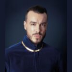 Performer Cem Adrian wearing a blue coat, against a dark blue background