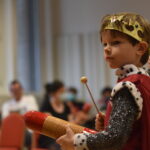 A small child in a medieval costume plays a small toy percussion instrument