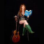Musician Kezia Gill in a sparkly outfit and green boots, holding a blue book and a guitar.