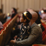 A young boy wearing a paper crown, sitting on a man's lap, watching a show