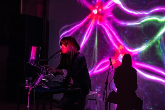 musician Helen Anahita Wilson on a dark stage with computer, lit with pink lights