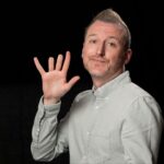 Comedian Stuart Mitchell, in a grey shirt, holding up his hand with the tip of his fourth finger missing