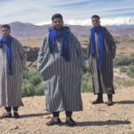 Three men in blue scarves and robes against a desert and mountain backdrop