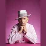 Musician Kevin Rowland wearing a pale pink suit and hat, on a dark pink background