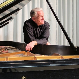 Craig Sheppard (a man with grey hair and a black shirt) leaning on a piano