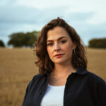 Image of singer Katherine Priddy, with brown curly hair wearing a white top and black jacket, in front of a wheat field