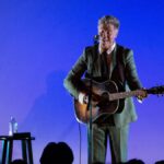 Musician Josh Ritter with guitar, wearing a grey jacket, against a blue background